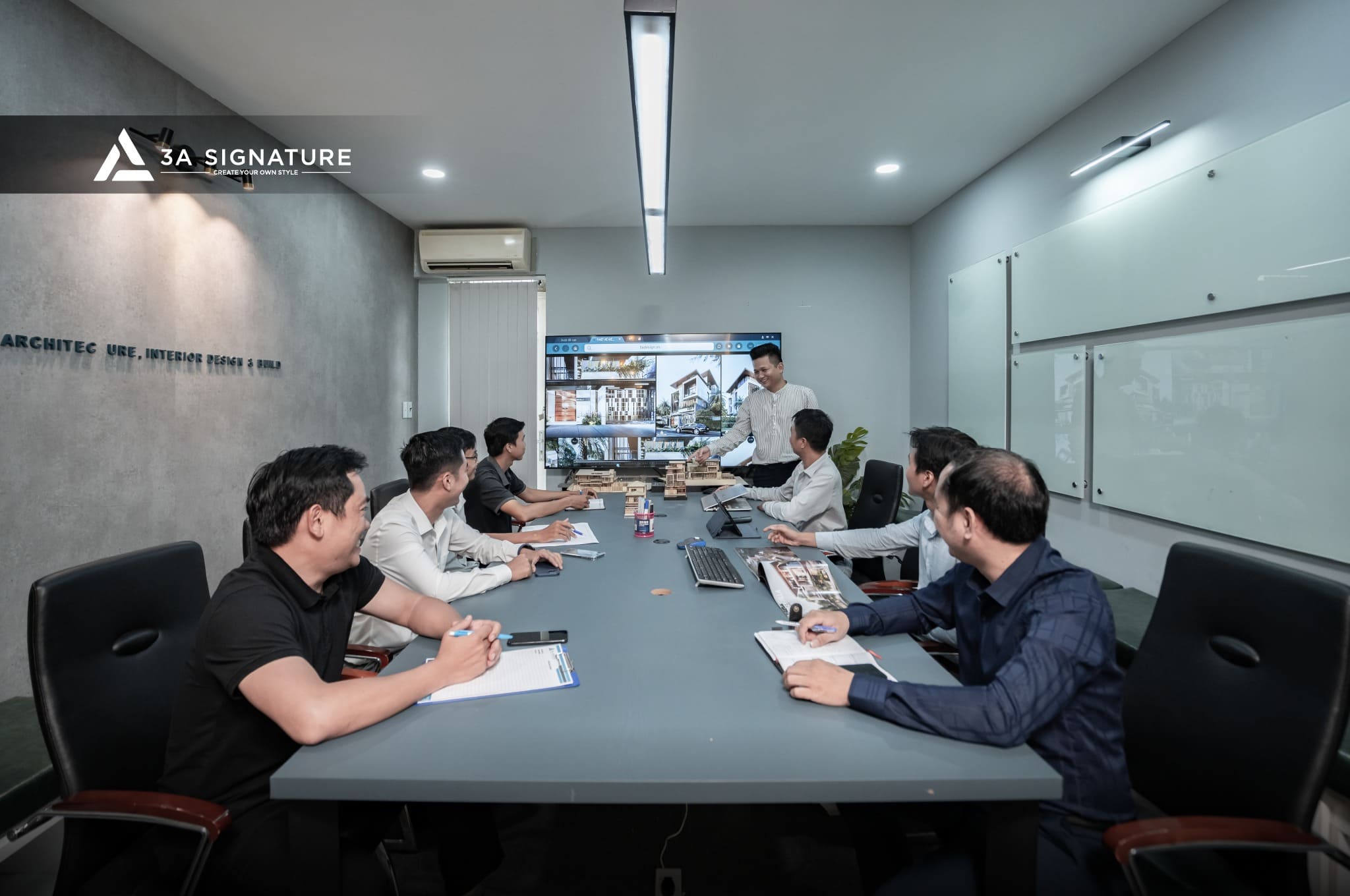 Dịch Vụ Thi Công Nhóm kiến trúc sư và lãnh đạo của 3A Signature đang họp trong văn phòng hiện đại, thuyết trình bản vẽ biệt thự Modern Tropical trên màn hình, thể hiện quy trình làm việc chuyên nghiệp và sáng tạo.
